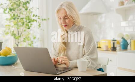 Femme âgée authentique utilisant un ordinateur portable dans une salle de cuisine lumineuse à la maison. Belle vieille femme Pensioner avec cheveux gris navigue sur Internet sur un Banque D'Images