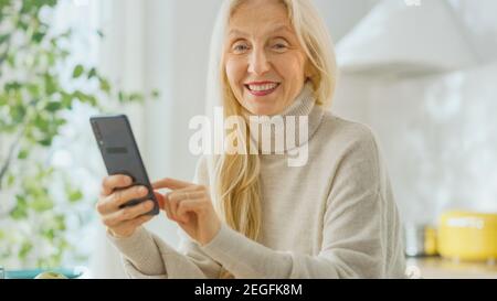 Femme âgée authentique utilisant un smartphone dans la cuisine à la maison. Belle ancienne femme Pensioner avec cheveux gris utilise une application sur un téléphone portable Banque D'Images