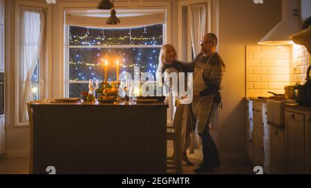 Heureux couple Senior en amour ont une soirée romantique, dansant dans la cuisine, célébrant l'anniversaire. Soirée romantique avec vin, table festive à l'intérieur Banque D'Images