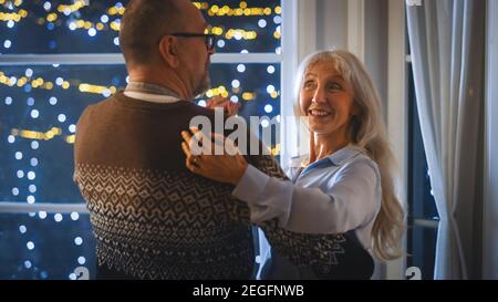 Heureux couple Senior en amour ont une soirée romantique, dansant dans la cuisine, célébrant l'anniversaire. Soirée romantique avec vin, table festive à l'intérieur Banque D'Images