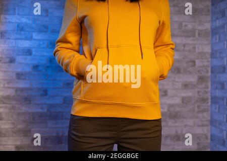 Gros plan des mains de la femme d'une personne méconnaissable dans des poches de sweat à capuche jaune sur fond de brique grise. Vue avant de la partie du corps de la jeune femme dans sw Banque D'Images