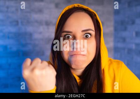 Portrait d'une jeune femme insatisfaite montrant le poing. Gros plan de la femelle affolée dans la cagoule jaune montre le poing, regardant l'appareil photo Banque D'Images