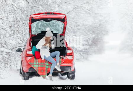 Femme dans des vêtements chauds assis dans le bois d'hiver tout en se penche sur la voiture et tenant une tasse de café. Banque D'Images