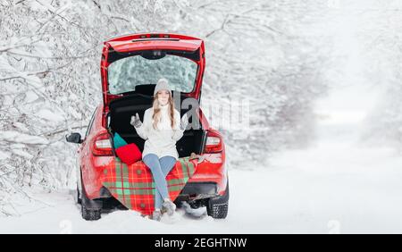 Une femme avec du café chaud dans ses mains est assise dans une voiture rouge lors d'une journée d'hiver enneigée dans la forêt. Banque D'Images