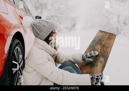 Une jeune femme s'assoit dans la neige au volant d'une voiture rouge et tient une carte entre ses mains. Concept de voyage. Banque D'Images