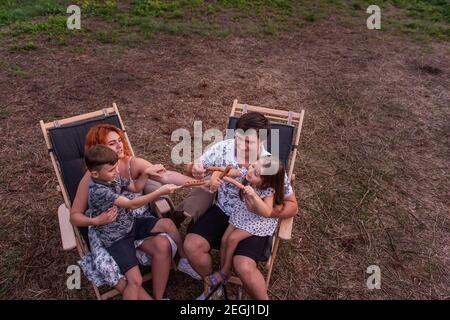 Une famille gaie pique-nique à l'extérieur de la ville, les saucisses sur brochettes sont frites au feu. Assis sur des chaises longues en bois près de la voie de la remorque. Les parents se trompaient Banque D'Images