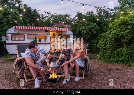 Une famille gaie pique-nique à l'extérieur de la ville, les saucisses sur brochettes sont frites au feu. Assis sur des chaises longues en bois près de la voie de la remorque. Les parents se trompaient Banque D'Images