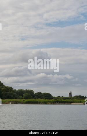 Une rive de rivière ou de lac, sous un ciel nuageux et détaillé.Lits reedbeds verts et arbres dans un bois.Un vieux moulin à vent abandonné (pompe à vent) dans la distance Banque D'Images