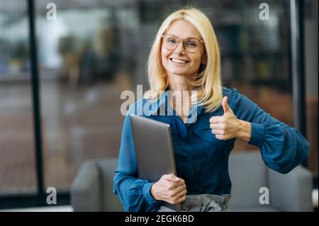 Belle femme d'affaires satisfaite posant au bureau, montrant les pouces vers le haut. Femme élégante en lunettes et supports de vêtements formels au bureau, regardant l'appareil photo, souriant Banque D'Images