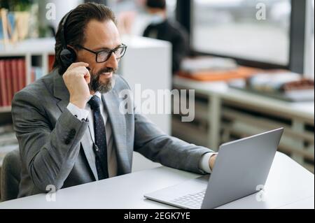 Un homme joyeux en matière de micro-casque et de lunettes regarde l'écran de l'ordinateur portable, ayant une réunion d'affaires en ligne. Un employé mûr discute de questions de travail avec ses collègues Banque D'Images