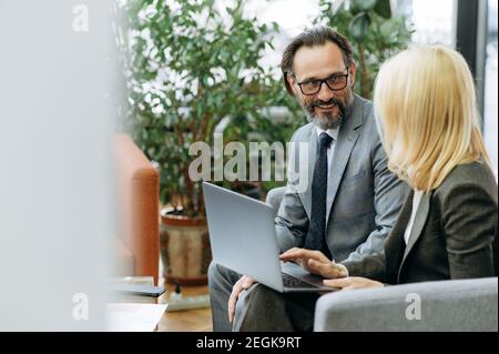Des collègues d'âge moyen prospères assis sur le canapé au bureau, discutant d'un nouveau plan d'affaires. Beau homme avec la femme blonde-cheveux utilisant l'ordinateur, parlant des idées, concept de partenariat Banque D'Images