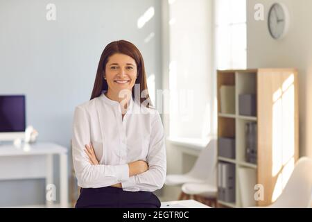 Bonne femme d'affaires en blouse blanche debout bras pliés dans le bureau et sourire à l'appareil photo Banque D'Images