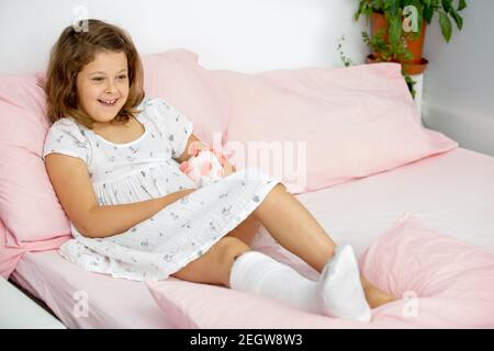 Une jeune fille joyeuse se trouve sur le lit dans un casting sur sa jambe, avec une cheville cassée. Jambe cassée chez un enfant. Banque D'Images