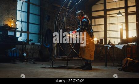 Talentueux artiste féminin Tomboy innovant utilise un meuleuse d'angle pour faire une sculpture de métal abstraite, brutale et expansive dans un atelier. Contemporain Banque D'Images