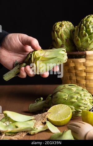 Vue de la main d'une femme tenant un artichaut pelé avec des artichauts dans un panier et des limes sur une table en bois, portrait Banque D'Images