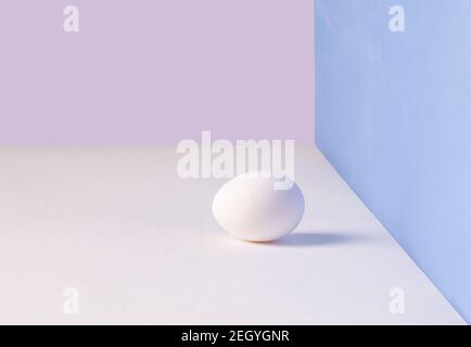 Œuf simple blanc. Œuf de poulet aux ombres douces sur une table blanche. Fond rose, bleu. Modèle pour les vacances de Pâques. Banque D'Images