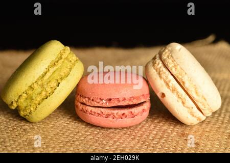 Trois macarons doux et savoureux, entièrement parfumés, reposent sur un tissu jute, sur un fond noir. Banque D'Images