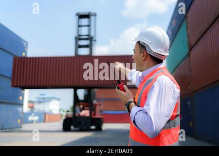 Le contremaître caucasien utilise talkie walkie pour commander ses travailleurs avec une grue de levage de conteneur en arrière-plan dans le terminal de dépôt de conteneur. Banque D'Images