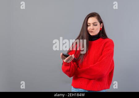 une jeune adolescente vêtue d'un chandail rouge regarde soigneusement son smartphone, isolé sur le gris Banque D'Images