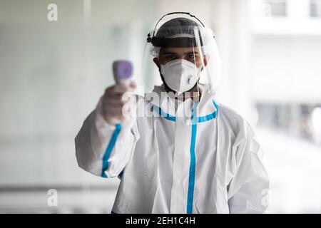 Portrait de l'homme médic portant un uniforme de protection biologique costume habillement, masque, gants mesurant la température corporelle avec thermomètre frontal infrarouge Banque D'Images