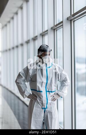 Un médecin de sexe masculin en costume de protection et un masque médical se trouve près d'une fenêtre dans un hôpital. Banque D'Images
