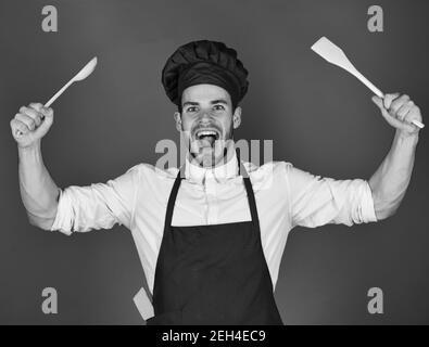 Le chef au visage excité tient une cuillère en bois et une spatule sur fond rouge. Ustensiles de cuisine et concept de cuisine. Homme en chapeau bordeaux et tablier avec outils de cuisson. Cuisine. Banque D'Images