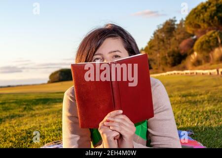 Une femme d'âge moyen se faisant un pelage sur un livre qu'elle tient, tout en étant couchée sur un peu d'herbe. Coucher de soleil crémeux en arrière-plan. Banque D'Images