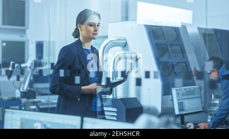 Usine moderne : une femme ingénieure confiante parcourt les installations, tient son ordinateur portable, inspecte et surveille le fonctionnement de l'atelier. Le fabricant a Banque D'Images