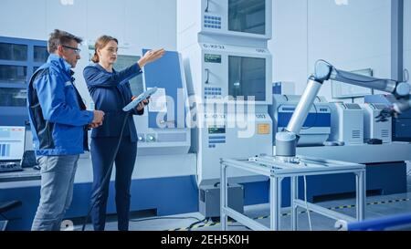 Modern Factory : responsable de projet féminin et ingénieur masculin debout, parlant et utilisant un Tablet PC pour programmer la ligne d'assemblage de bras de robot Banque D'Images