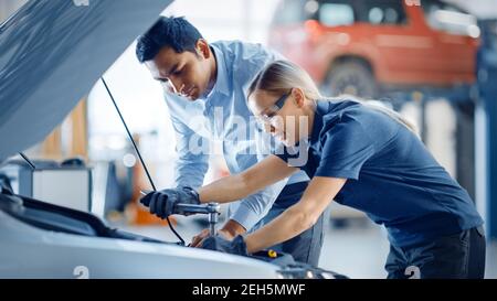 Le responsable vérifie les résultats des diagnostics sur une tablette et explique une panne de moteur à un mécanicien féminin. L'employé du service automobile utilise une clé à cliquet pour Banque D'Images