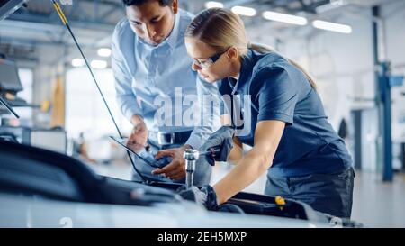 Le responsable vérifie les résultats des diagnostics sur une tablette et explique une panne de moteur à un mécanicien féminin. L'employé du service automobile utilise une clé à cliquet pour Banque D'Images