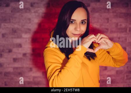 Portrait de la jeune femme montrant le signe du coeur avec ses mains. Gros plan de Happy brunette montre le signe de l'amour sur fond rétroéclairé Banque D'Images
