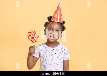 Gros plan de petite adorable fille afro-américaine dans le chapeau de partie tenant le lollipop doux dans les mains, posant à l'appareil photo sur fond jaune, Copy Space Banque D'Images