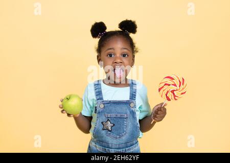 Une alimentation saine, des aliments appropriés, des bonbons et des fruits. Drôle adorable africaine petite fille tient le Lollipop coloré et la pomme verte et montre sa langue à Banque D'Images