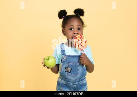 Petite mignonne jolie fille africaine mangeant un bonbon Lollipop tout en tenant la pomme verte dans l'autre main, isolé sur fond jaune. Choix entre Banque D'Images