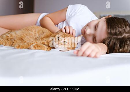 Une fille avec un chat de gingembre se trouve sur un lit blanc. Une jeune fille est à la course d'un chat au gingembre en étant couché sur une feuille blanche Banque D'Images