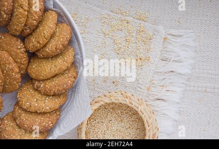 Biscuits de sésame sur une assiette blanche, graines de sésame dans un panier en osier, nappe tissée rugueuse, plat, espace de copie Banque D'Images