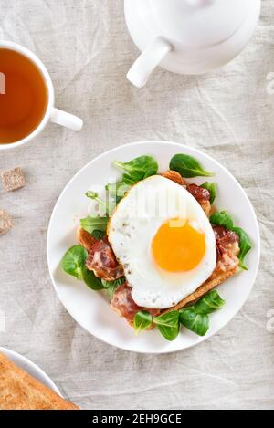 Des sandwichs ouverts avec des œufs frits, du bacon et des feuilles de légumes sur une assiette sur fond clair. Vue de dessus, plan d'appartement Banque D'Images