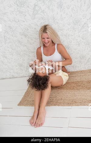 Portrait de la jeune blonde blanche mère tenue joue tickles petite fille afro-américaine dans les bras. La fille rit se trouve sur les jambes de la femme. Tenderne Banque D'Images