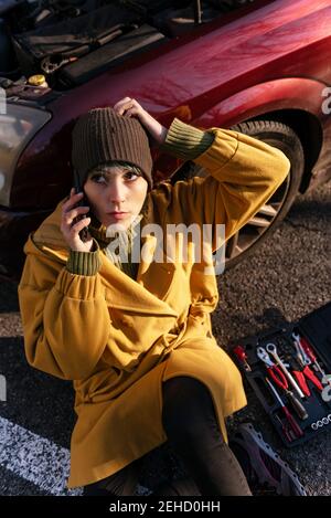 Calme jeune femme dans des vêtements chauds ayant un appel téléphonique et assis sur l'asphalte, assis sur la voiture avec le capot ouvert près de la boîte à outils sur le bord de la route et en regardant un Banque D'Images