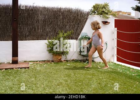 Vue latérale de la petite fille mignonne en maillot de bain marche et arrosage de la pelouse verte par le tuyau pendant les vacances d'été dans la campagne Banque D'Images