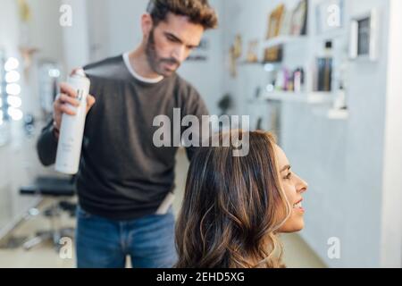 Vue latérale d'une jeune femme jeune et heureuse qui souriait pendant styliste masculine concentrée appliquant un spray capillaire dans un salon de beauté moderne Banque D'Images