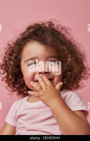 Adorable petite fille gaie aux cheveux bouclés dans des vêtements décontractés couvrir la bouche avec la main tout en regardant loin en souriant sur le rose arrière-plan Banque D'Images