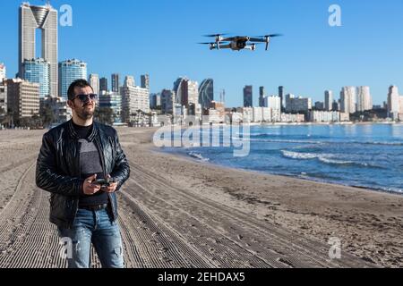 Homme sérieux dans des lunettes de soleil avec télécommande debout sur la plage drone de vol sur la côte près de la ville Banque D'Images