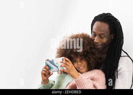Père américain africain positif embrassant la fille avec la coiffure d'Afro pendant partager des vidéos amusantes sur un téléphone portable Banque D'Images