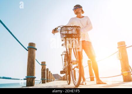 Portrait d'un homme heureux souriant pieds nus vêtu de vêtements d'été légers et de lunettes de soleil à vélo sur la jetée en bois. Des vacances imprudentes Banque D'Images