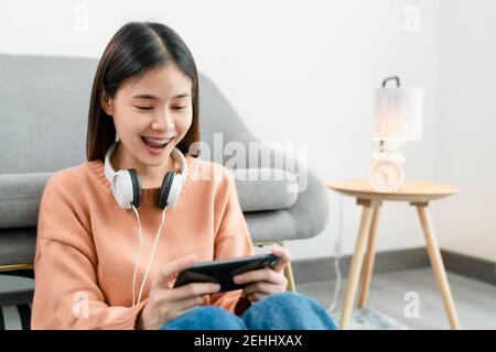 Une jeune femme asiatique excitée porte un casque blanc sur la tête et joue à des jeux sur smartphone dans le salon de la maison. Banque D'Images