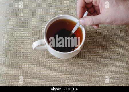 La main d'un homme mélange le thé noir dans une tasse avec une cuillère en métal. Un mug en céramique blanc rempli de thé noir. Les feuilles de thé infusé se trouvent au fond du Banque D'Images