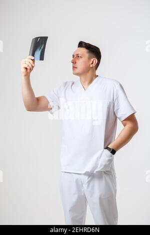 Un jeune médecin masculin dans un costume chirurgical blanc tient et Examine une image radiographique des os de la jambe du patient. Isolé sur fond blanc Banque D'Images