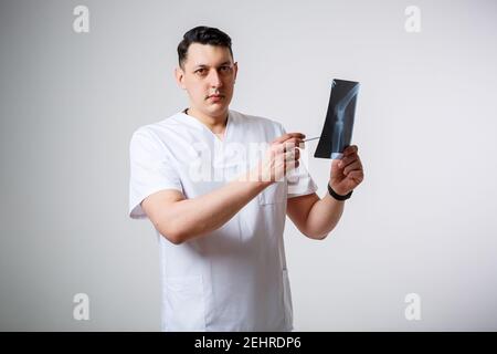 Un jeune médecin masculin dans un costume chirurgical blanc tient et Examine une image radiographique des os de la jambe du patient. Isolé sur fond blanc Banque D'Images